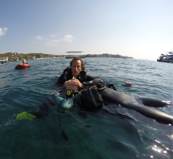 Instructeur en combinaison aidant un plongeur flottant à la surface près de Nusa Lembongan, mer calme et bateaux en arrière‑plan