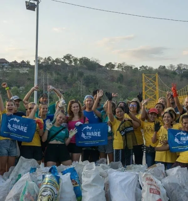 Bénévoles à Nusa Lembongan souriant et levant les mains après une collecte de déchets, sacs remplis de détritus empilés devant eux.