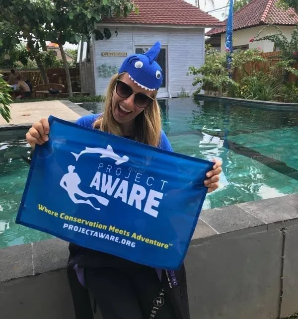 Femme souriante à Nusa Lembongan tenant une bannière bleu "Project Aware" près d'une piscine, portant un bonnet requin et des lunettes de soleil.